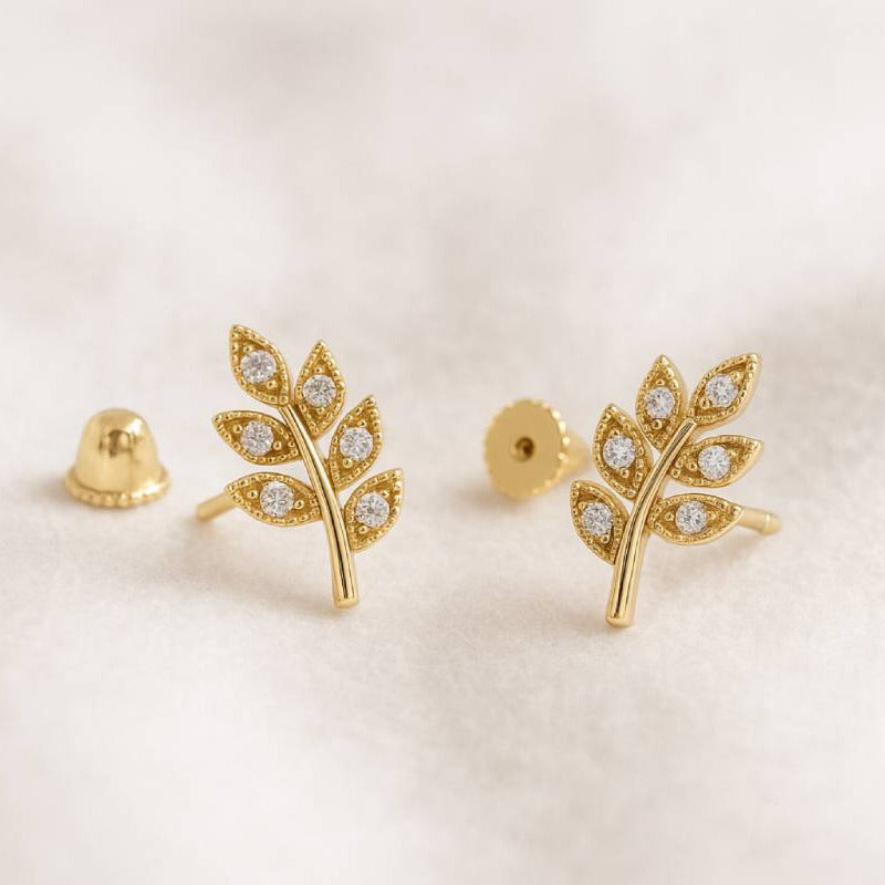 Gold leaf-shaped earrings with embedded stones on a light beige background