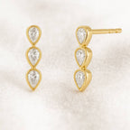 Gold earrings with three clear gemstones on a light beige background